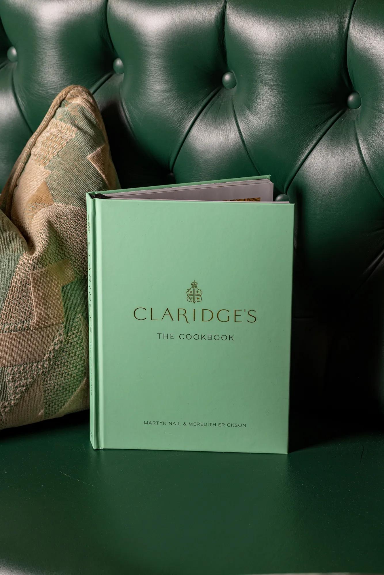 Mint green hardback copy of Claridge’s: The Cookbook standing on a deep green tufted leather banquette beside a patterned cushion.