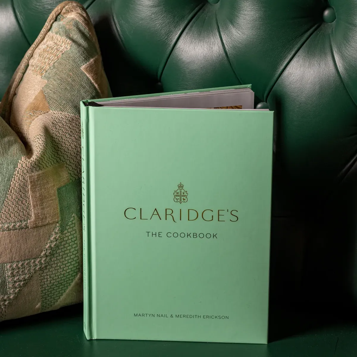 Mint green hardback copy of Claridge’s: The Cookbook standing on a deep green tufted leather banquette beside a patterned cushion.
