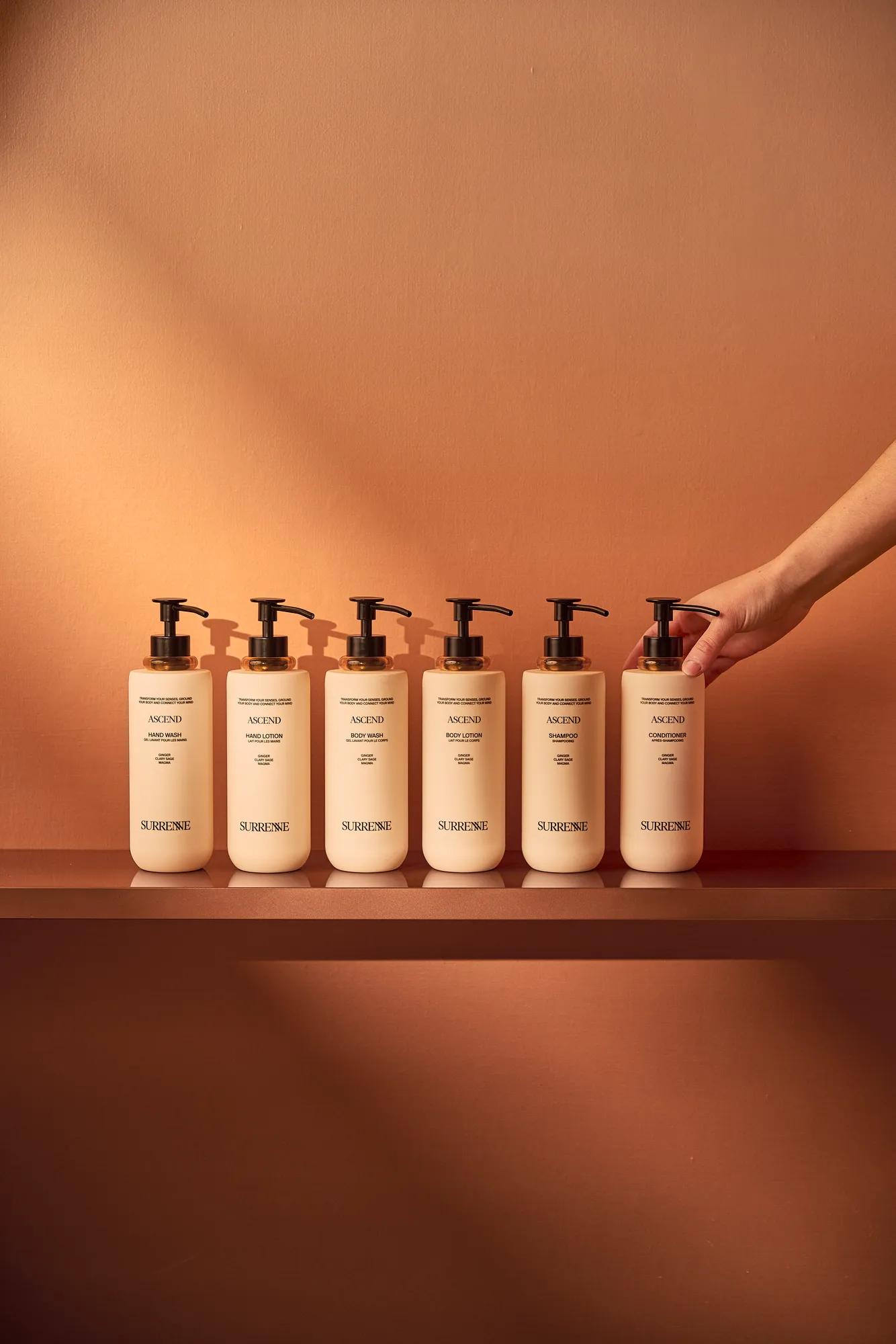 Six Surrenne Ascend bath and body bottles displayed on a shelf against a warm terracotta background.