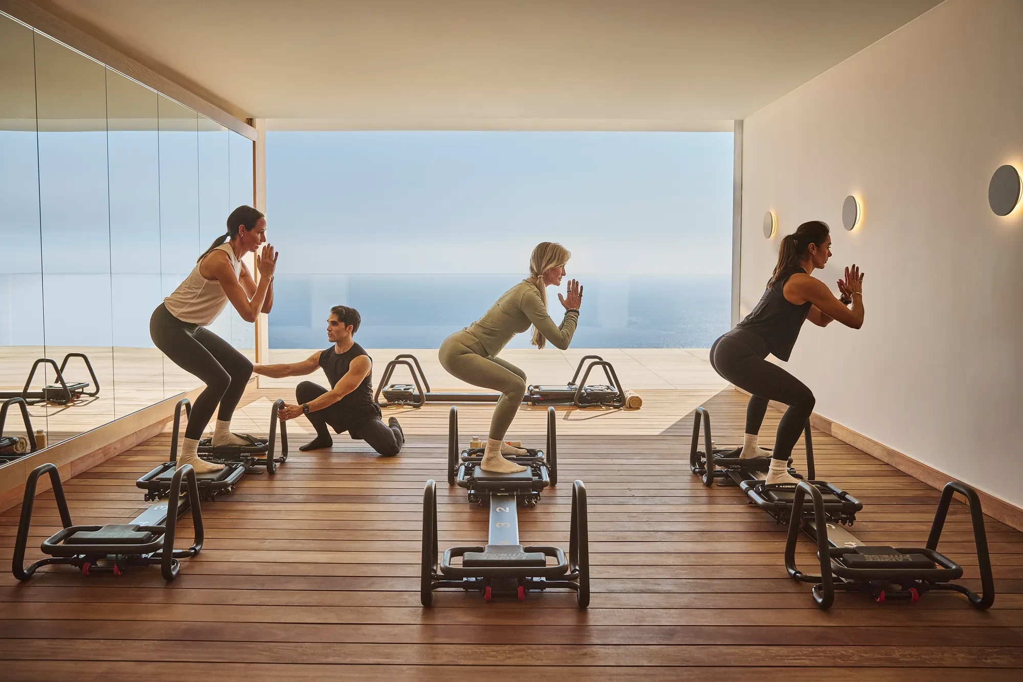 Group exercising on reformer machines in a fitness studio with floor-to-ceiling windows overlooking the sea.