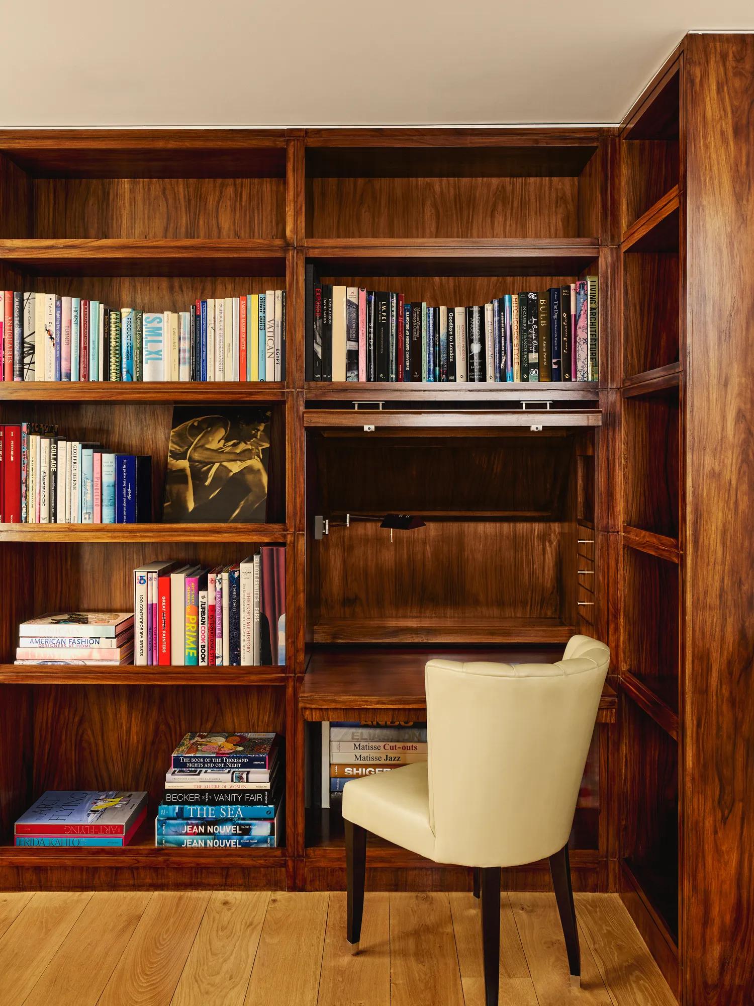 Warm wood library nook with shelves of colorful books and a cream upholstered chair at a small built-in desk.