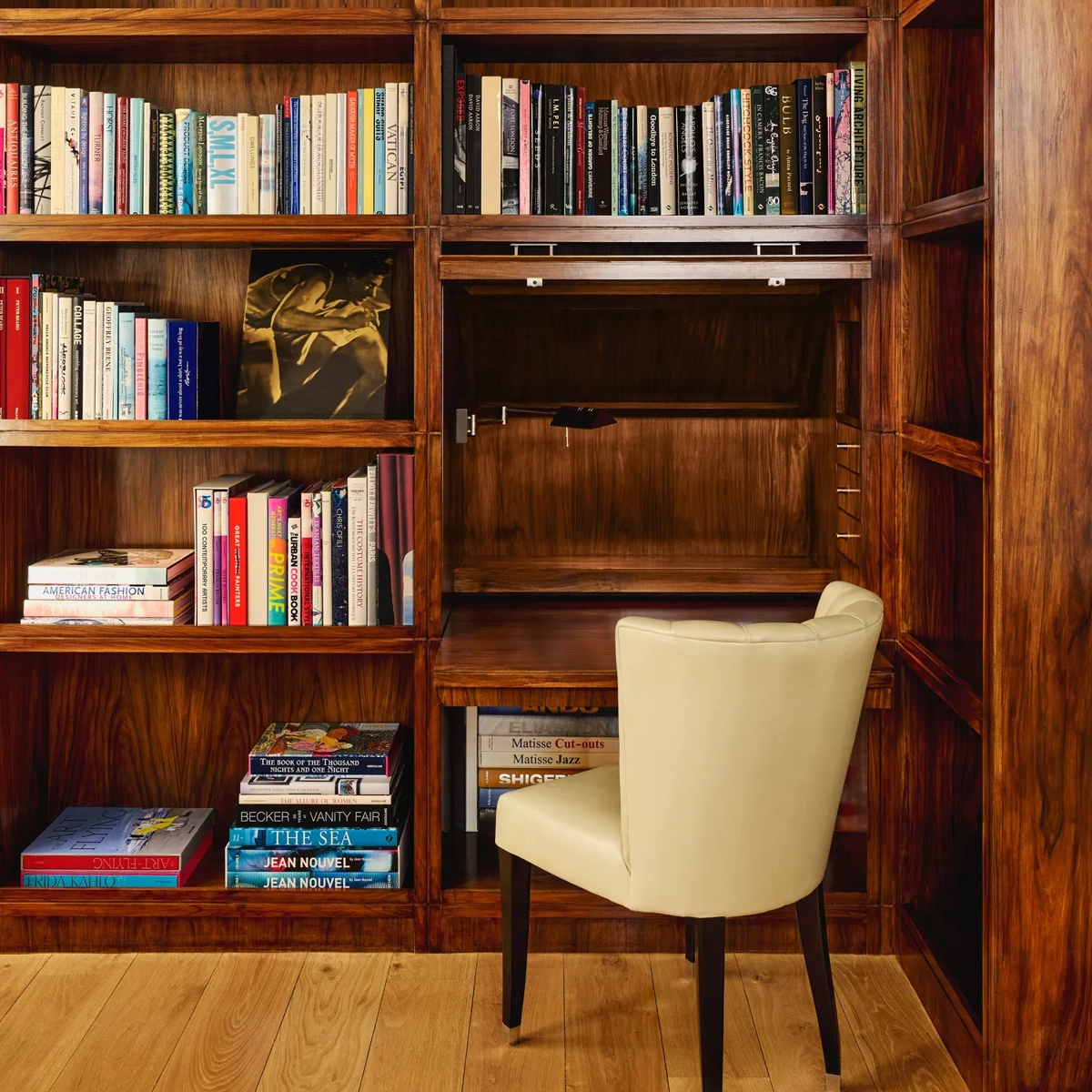 Warm wood library nook with shelves of colorful books and a cream upholstered chair at a small built-in desk.