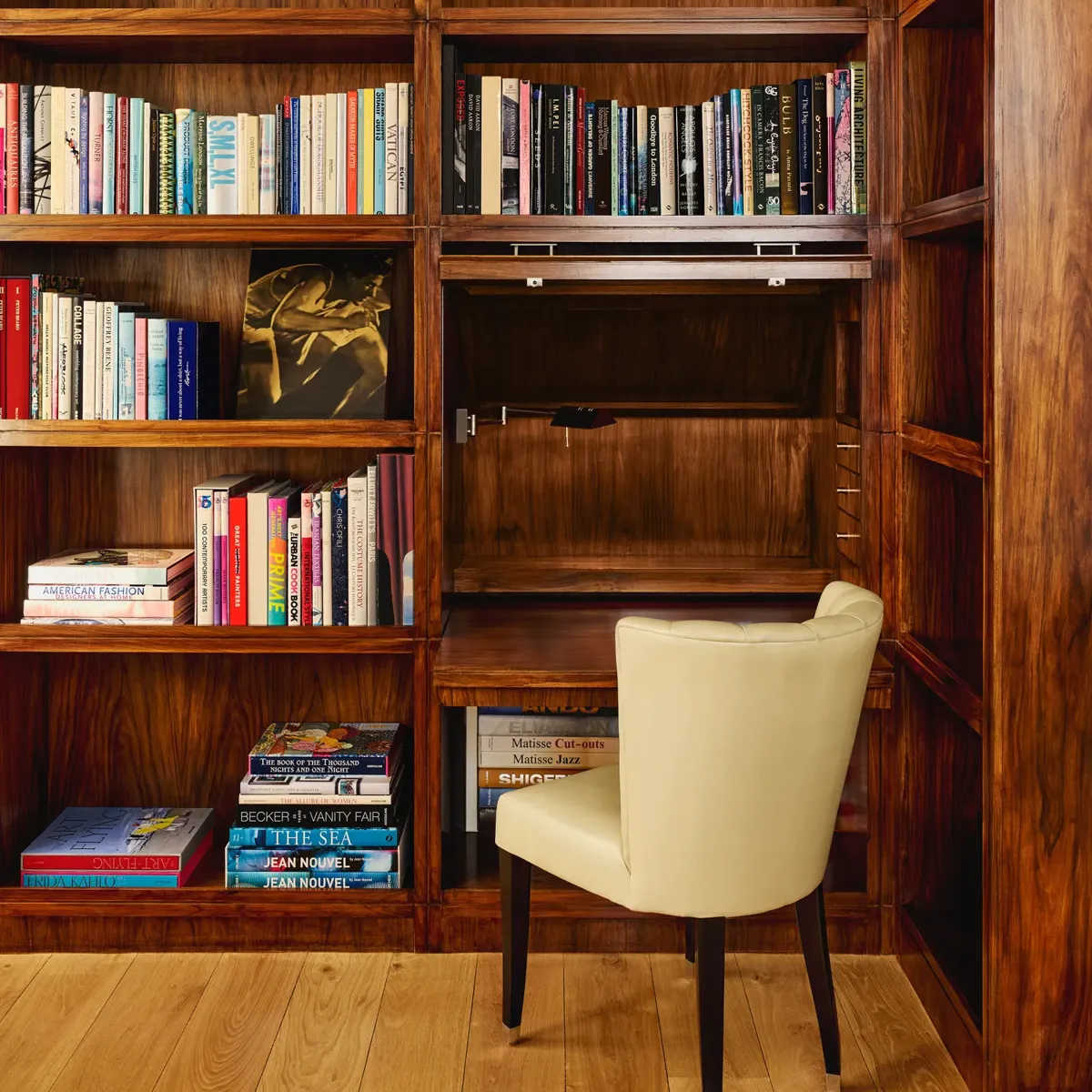 Warm wood library nook with shelves of colorful books and a cream upholstered chair at a small built-in desk.