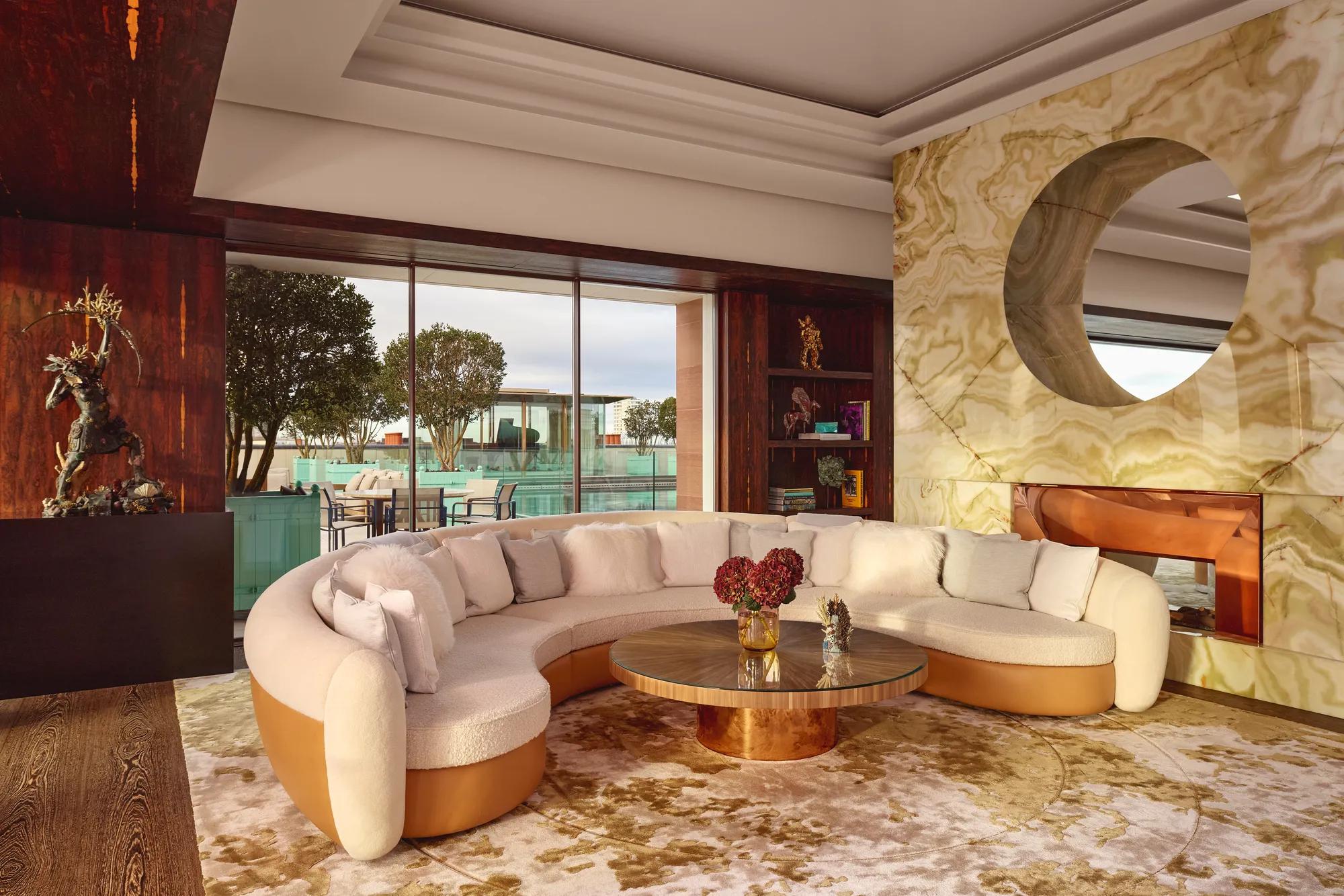 Spacious penthouse living area at Claridge’s with a curved cream sofa, marble fireplace, and glass doors opening to a terrace.