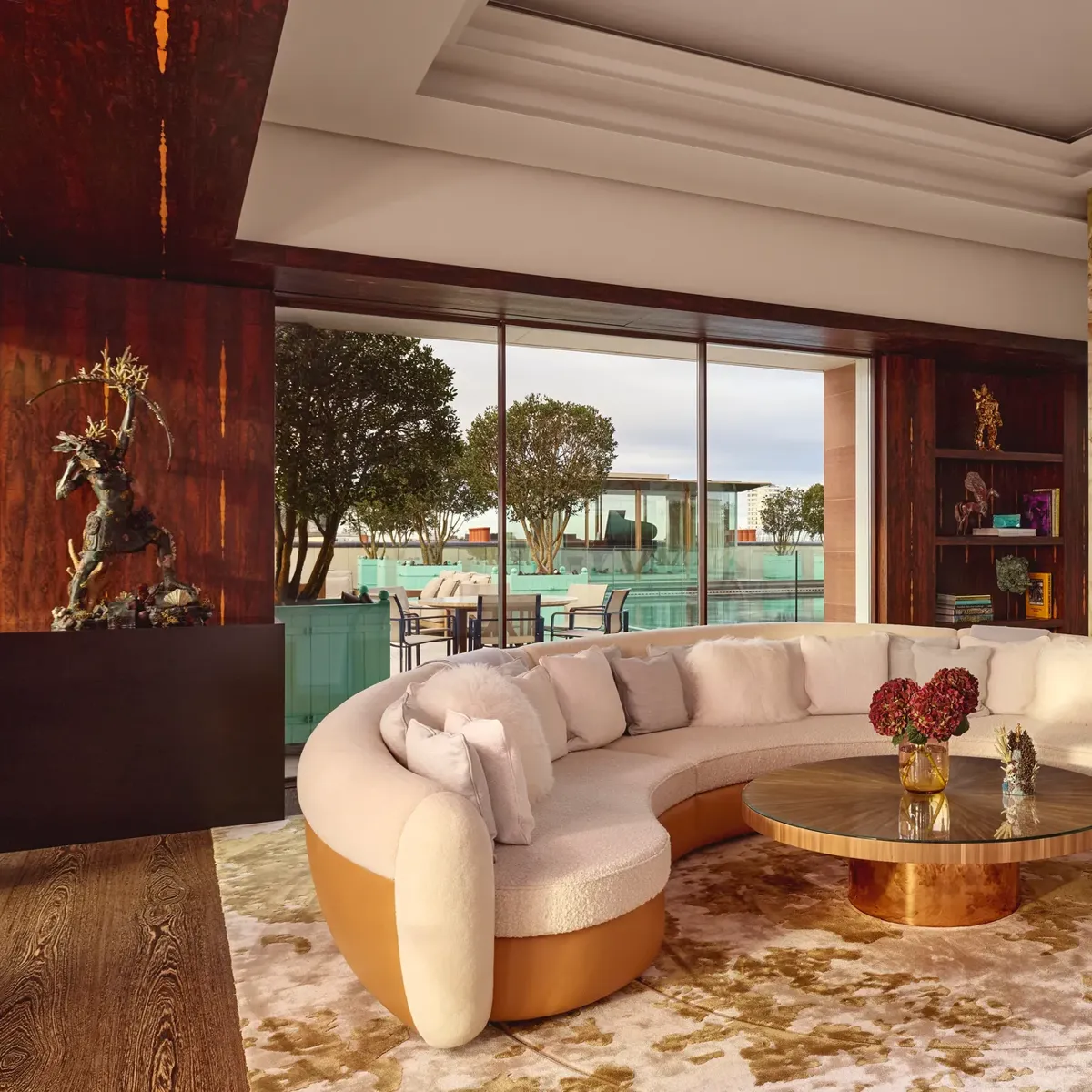 Spacious penthouse living area at Claridge’s with a curved cream sofa, marble fireplace, and glass doors opening to a terrace.