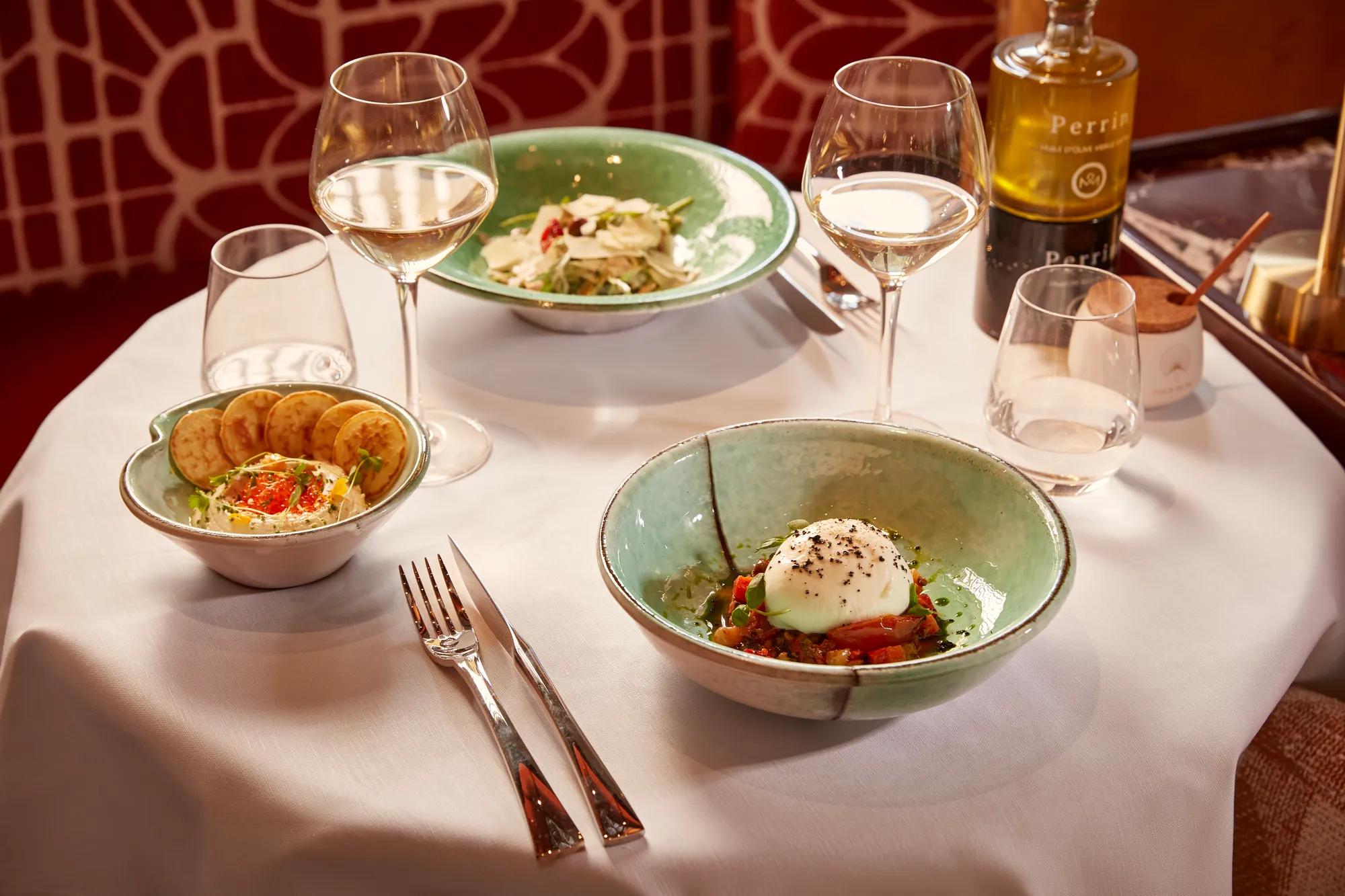 Table with three green ceramic bowls of appetisers, white wine glasses, and olive oil bottle.