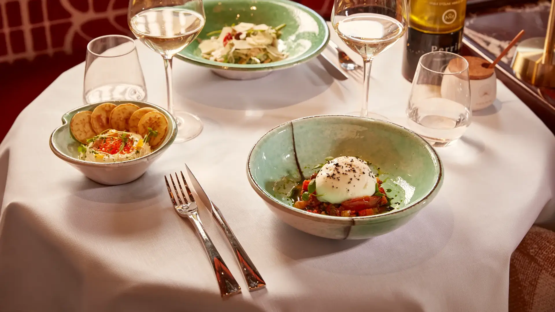 Table with three green ceramic bowls of appetisers, white wine glasses, and olive oil bottle.