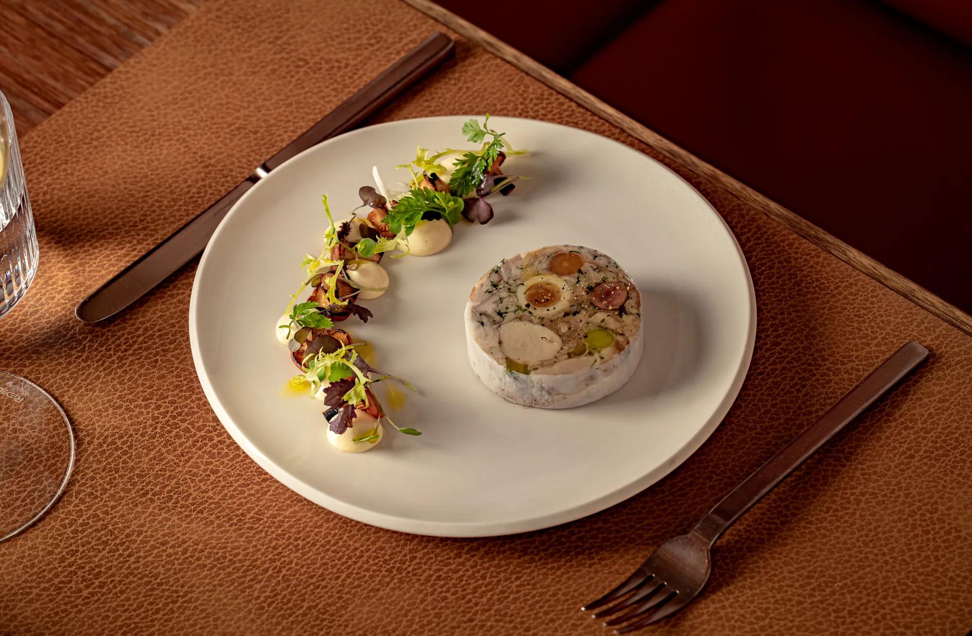 Delicately plated dish featuring a circular terrine with vegetables, served alongside artfully arranged greens and dollops of sauce on a white plate.