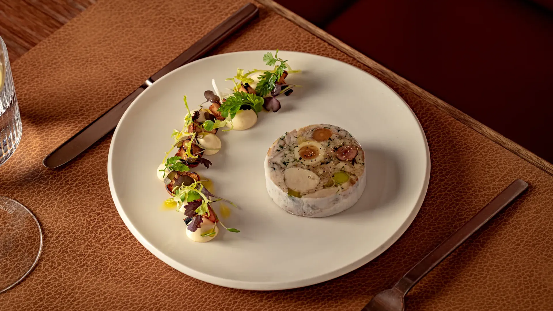 Delicately plated dish featuring a circular terrine with vegetables, served alongside artfully arranged greens and dollops of sauce on a white plate.