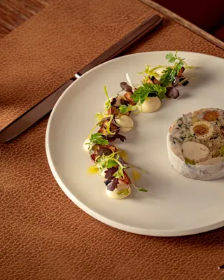 Delicately plated dish featuring a circular terrine with vegetables, served alongside artfully arranged greens and dollops of sauce on a white plate.