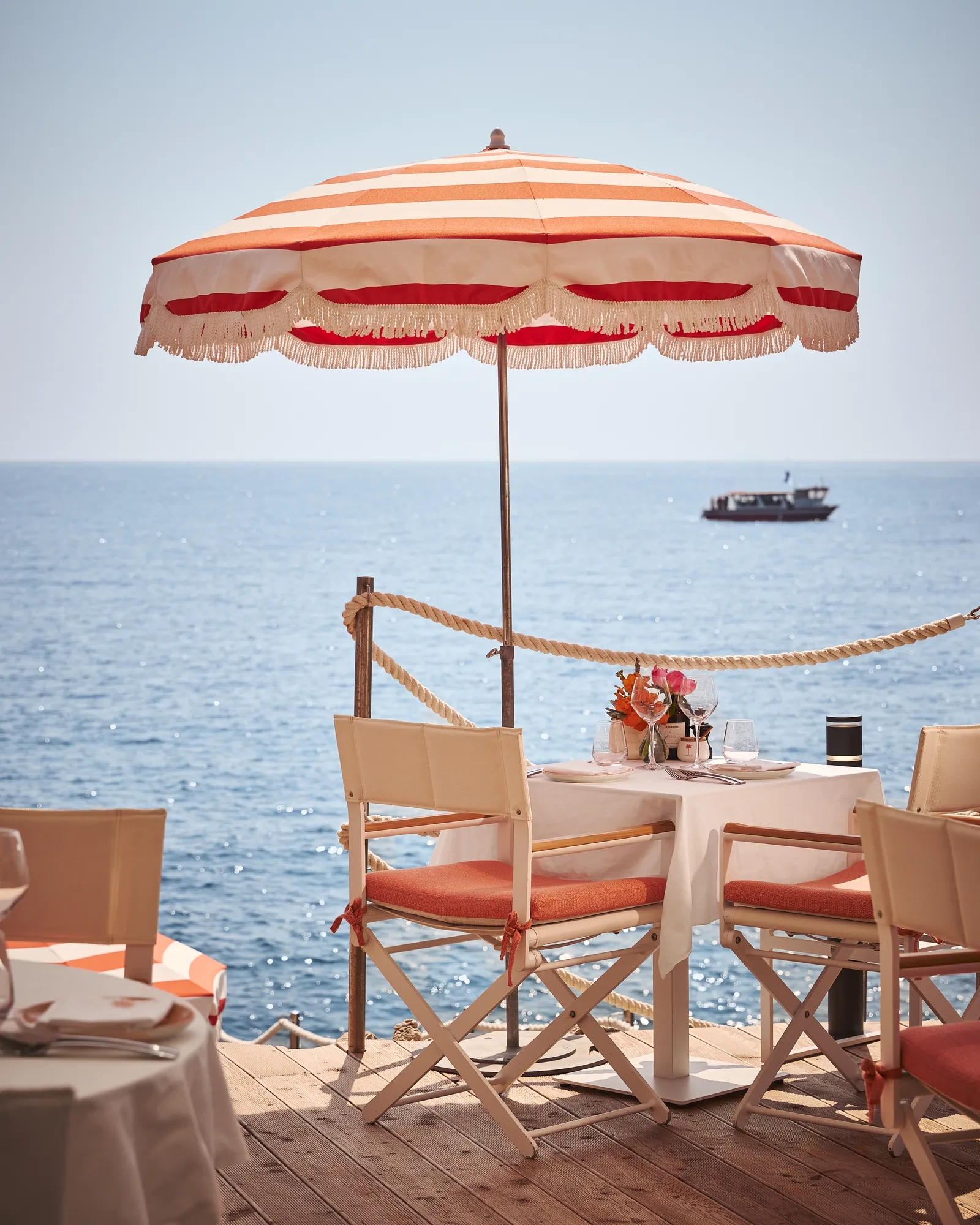 Seaside dining table with coral chairs and flowers beneath a red-striped umbrella, overlooking the ocean with passing boat