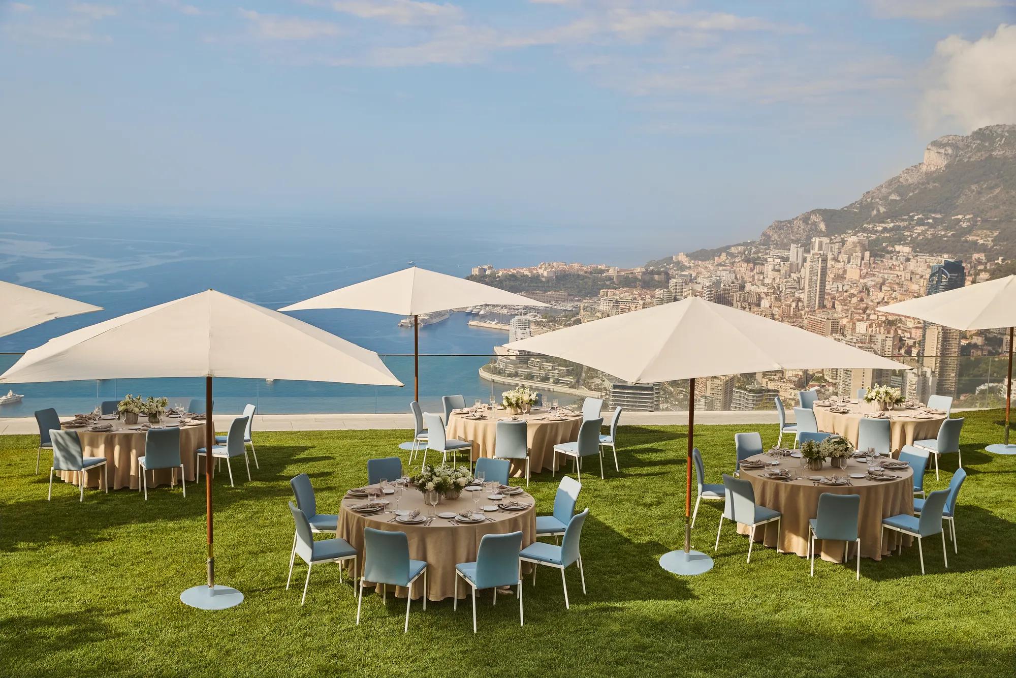 Outdoor dining setup with round tables, umbrellas, and sea views overlooking Monaco’s coastline from a grassy terrace.