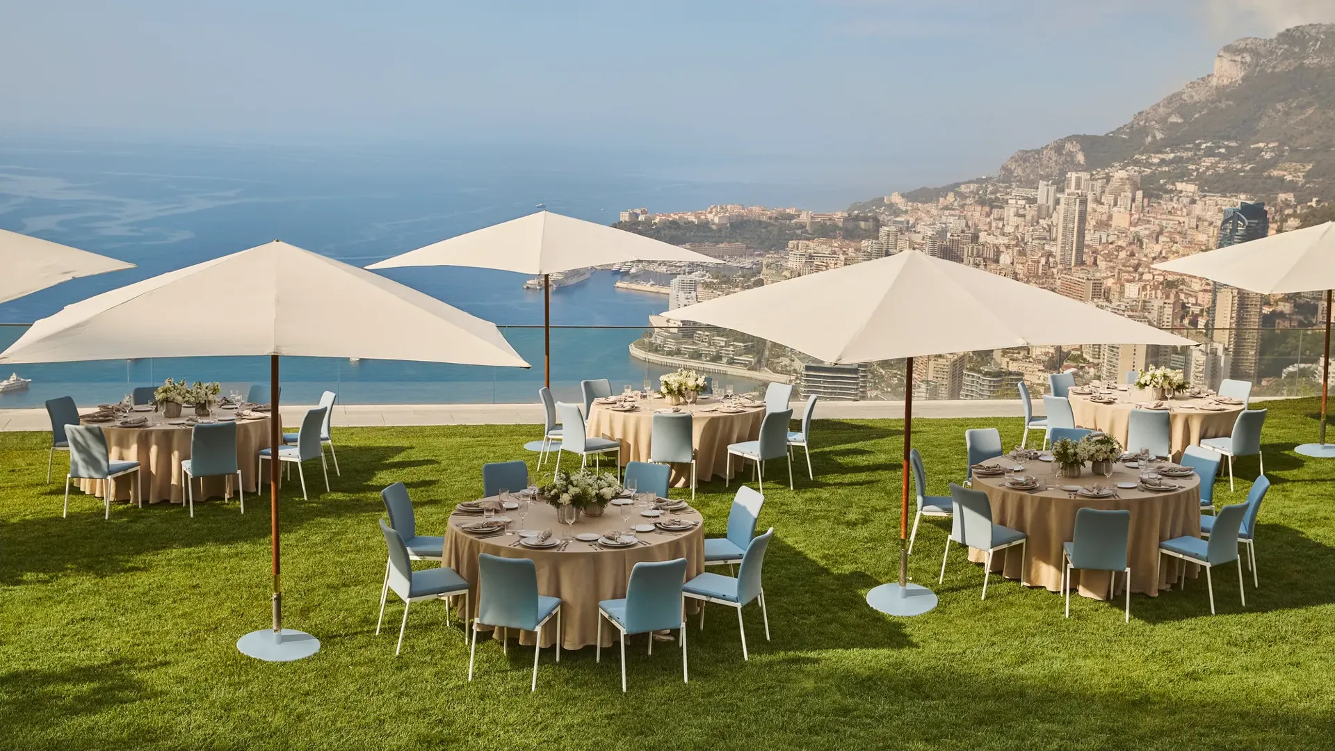 Outdoor dining setup with round tables, umbrellas, and sea views overlooking Monaco’s coastline from a grassy terrace.