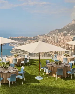 Outdoor dining setup with round tables, umbrellas, and sea views overlooking Monaco’s coastline from a grassy terrace.