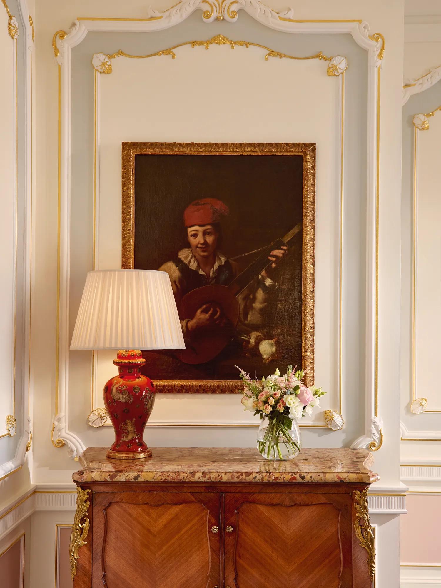 Ornate interior with antique cabinet, red table lamp, floral arrangement, and framed portrait painting.
