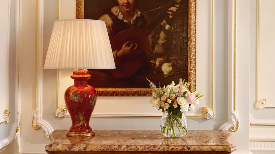 Ornate interior with antique cabinet, red table lamp, floral arrangement, and framed portrait painting.
