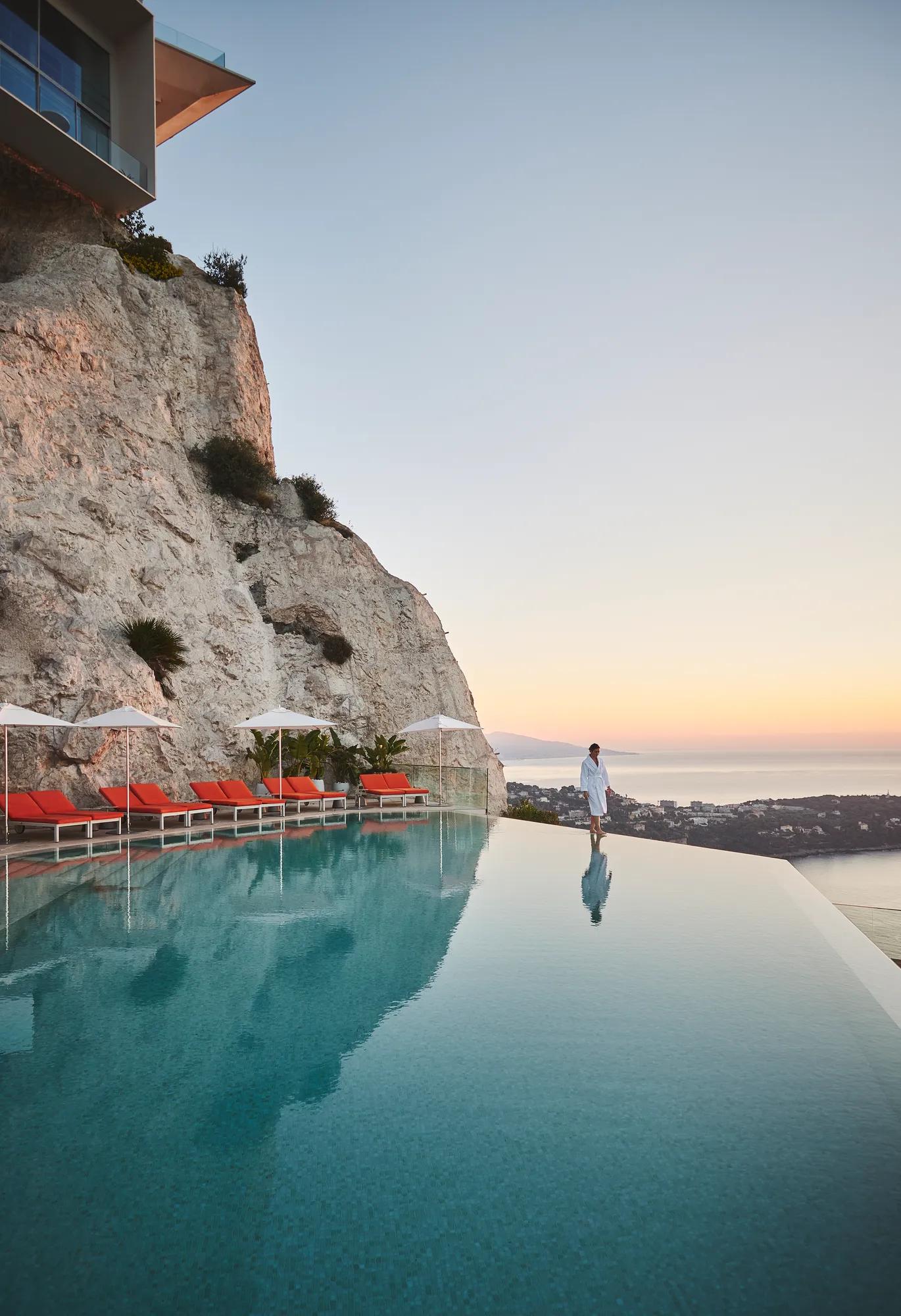 Infinity pool set against a cliffside with loungers and umbrellas, overlooking the sea at sunset with a guest walking along the edge