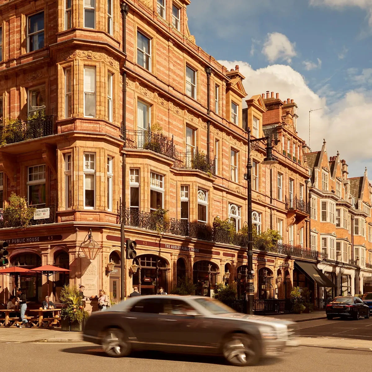 A sunlit street in Mayfair lined with red-brick Victorian buildings, including The Audley Public House with outdoor seating, as cars pass through the busy London intersection under a partly cloudy sky.