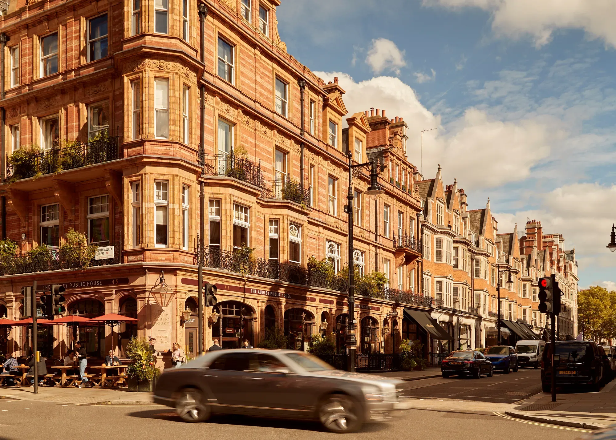 A sunlit street in Mayfair lined with red-brick Victorian buildings, including The Audley Public House with outdoor seating, as cars pass through the busy London intersection under a partly cloudy sky.