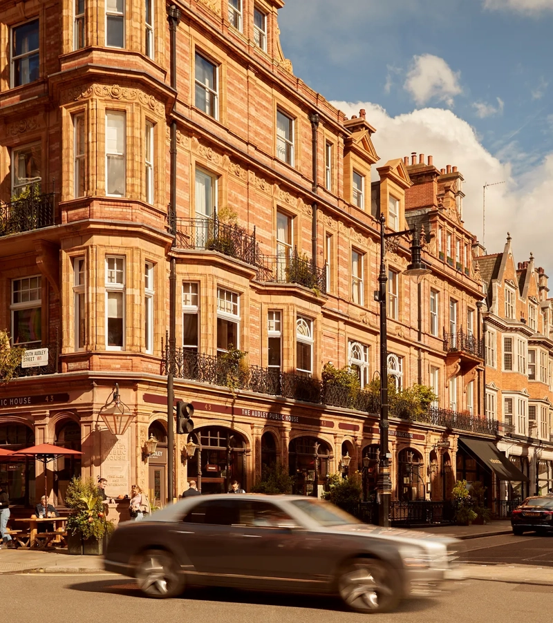 A sunlit street in Mayfair lined with red-brick Victorian buildings, including The Audley Public House with outdoor seating, as cars pass through the busy London intersection under a partly cloudy sky.