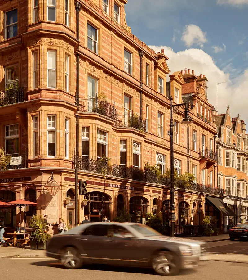A sunlit street in Mayfair lined with red-brick Victorian buildings, including The Audley Public House with outdoor seating, as cars pass through the busy London intersection under a partly cloudy sky.
