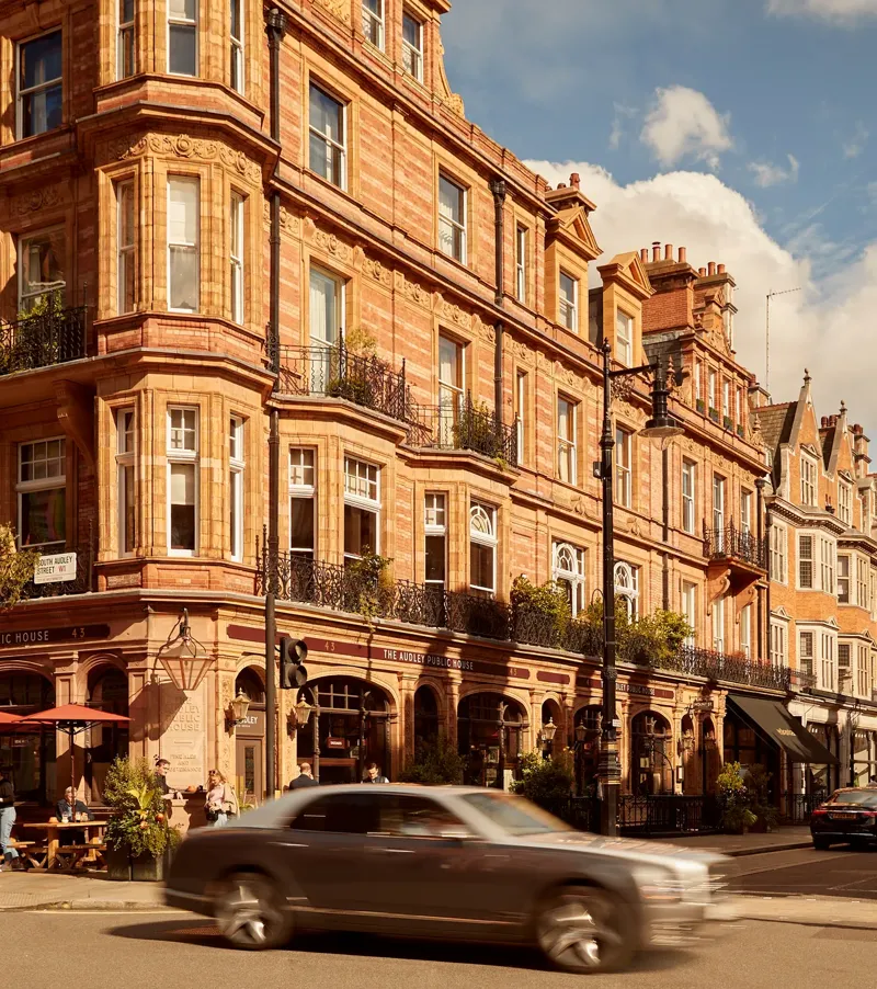 A sunlit street in Mayfair lined with red-brick Victorian buildings, including The Audley Public House with outdoor seating, as cars pass through the busy London intersection under a partly cloudy sky.