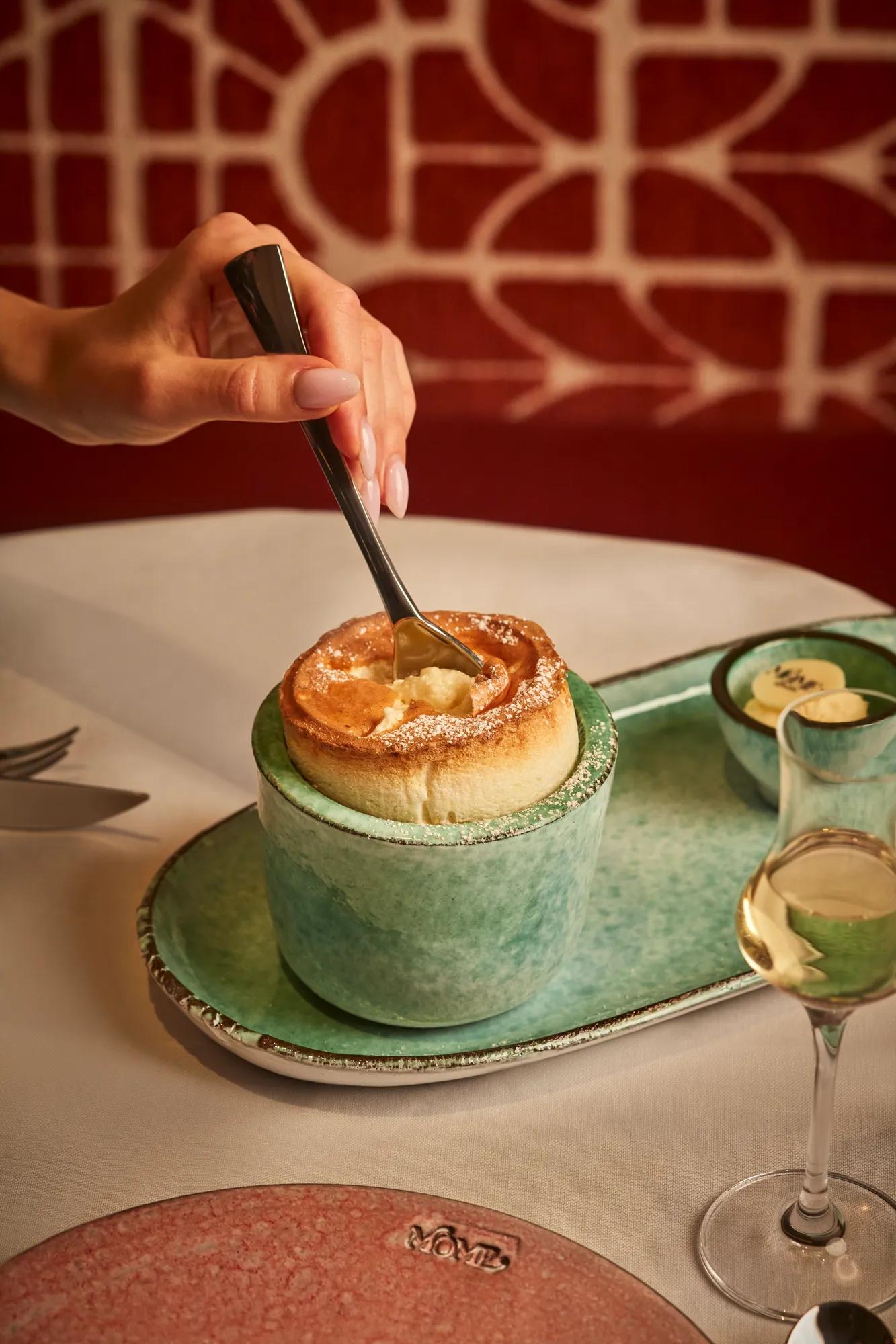 Hand dipping spoon into golden soufflé in green ceramic cup, with dessert wine glass on table.