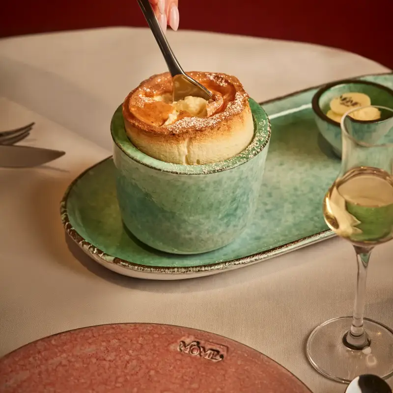 Hand dipping spoon into golden soufflé in green ceramic cup, with dessert wine glass on table.