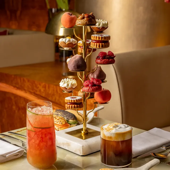 Tiered stand of assorted pastries with two cocktails served on a table in a refined lounge setting.