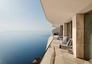 Balcon avec fauteuils rayés et transats derrière un garde-corps en verre, offrant une vaste vue sur la mer sous un ciel bleu clair.