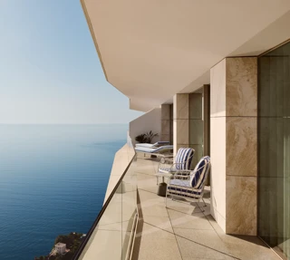 Balcony with striped chairs and loungers behind a glass railing, offering expansive sea views beneath a clear blue sky.