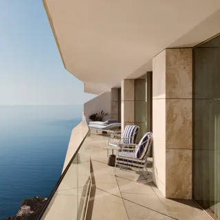 Balcon avec fauteuils rayés et transats derrière un garde-corps en verre, offrant une vaste vue sur la mer sous un ciel bleu clair.