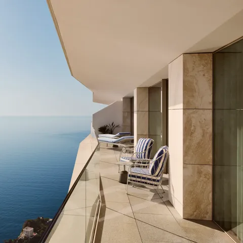 Balcony with striped chairs and loungers behind a glass railing, offering expansive sea views beneath a clear blue sky.