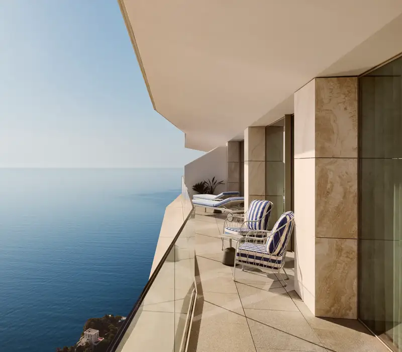 Balcony with striped chairs and loungers behind a glass railing, offering expansive sea views beneath a clear blue sky.