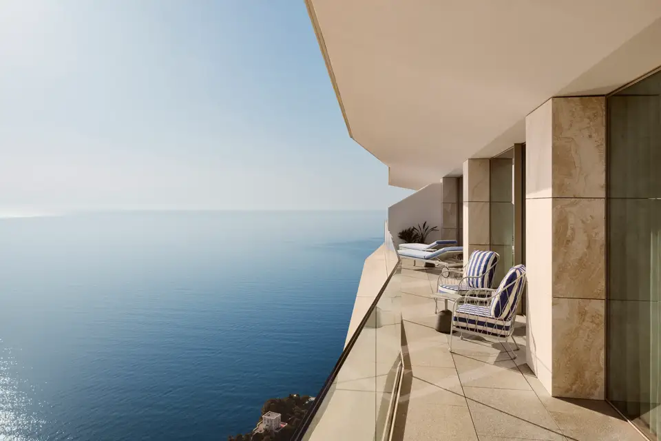 Balcony with striped chairs and loungers behind a glass railing, offering expansive sea views beneath a clear blue sky.