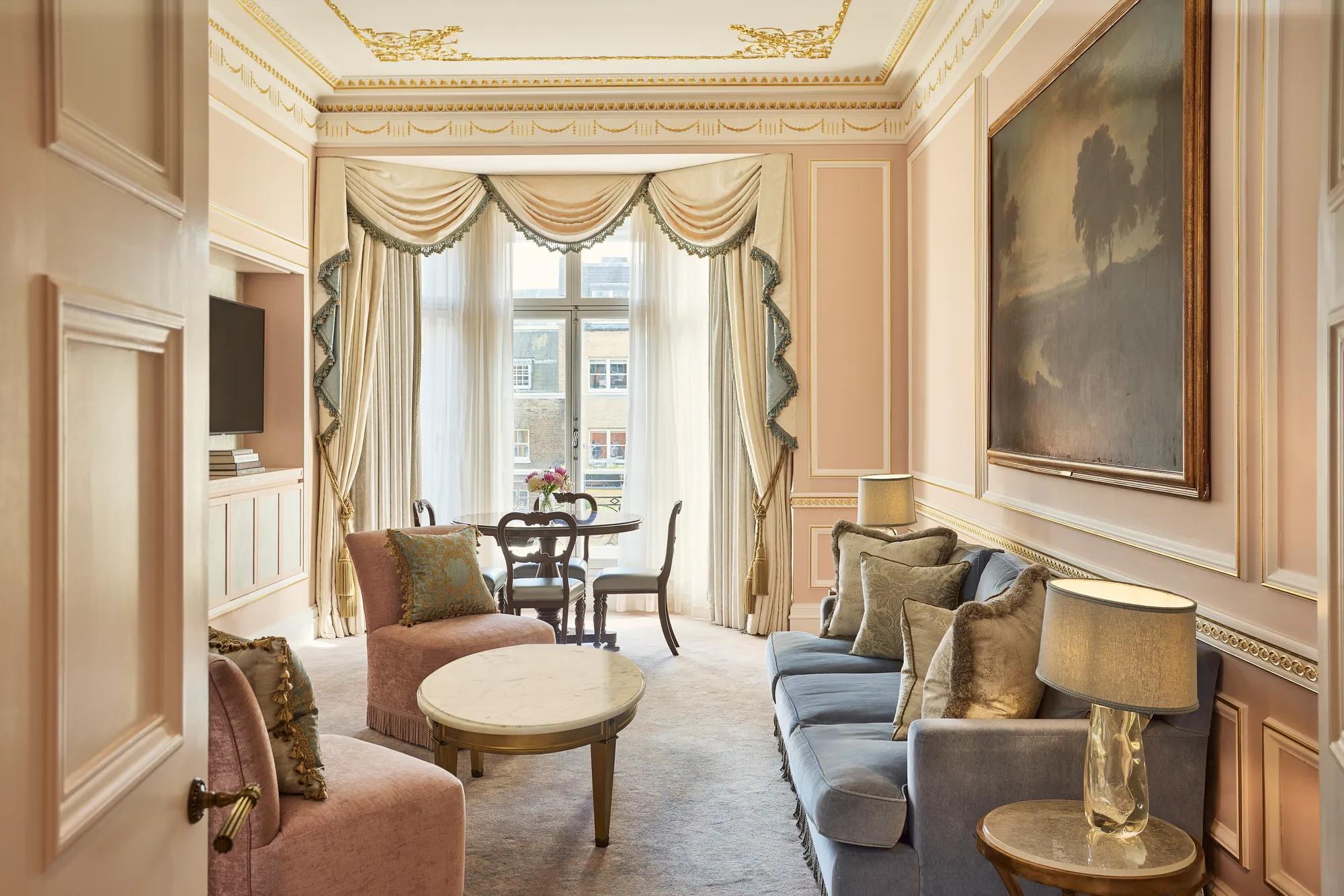 Elegant sitting room with blue and blush upholstered seating, a marble-topped coffee table, and a dining table positioned beside tall draped windows.