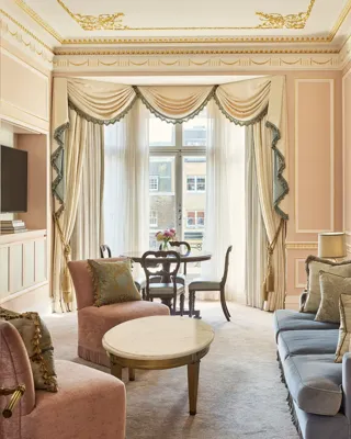Elegant sitting room with blue and blush upholstered seating, a marble-topped coffee table, and a dining table positioned beside tall draped windows.