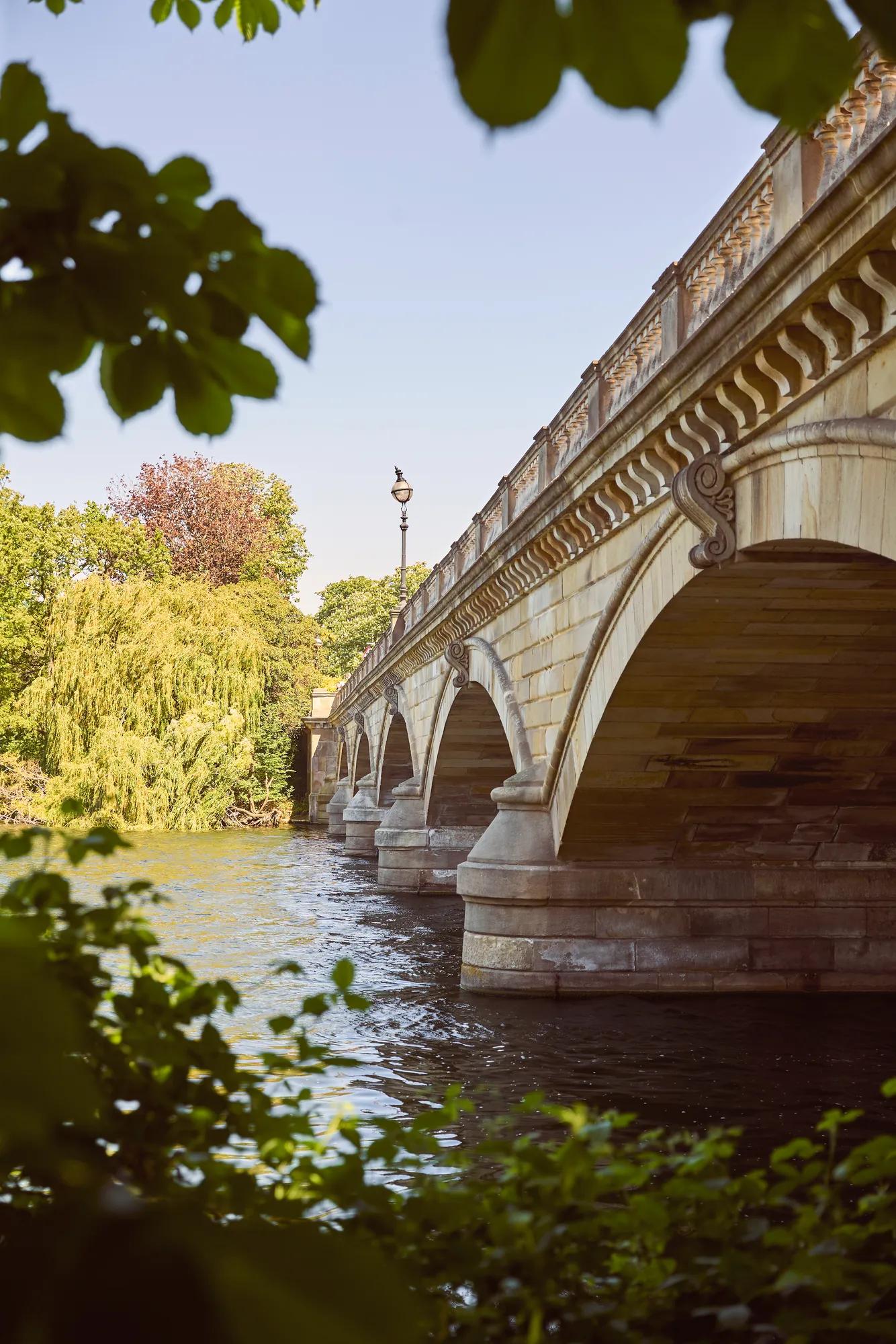 Stone arch bridge spanning a river, framed by leafy trees, with sunlight reflecting on the water below.