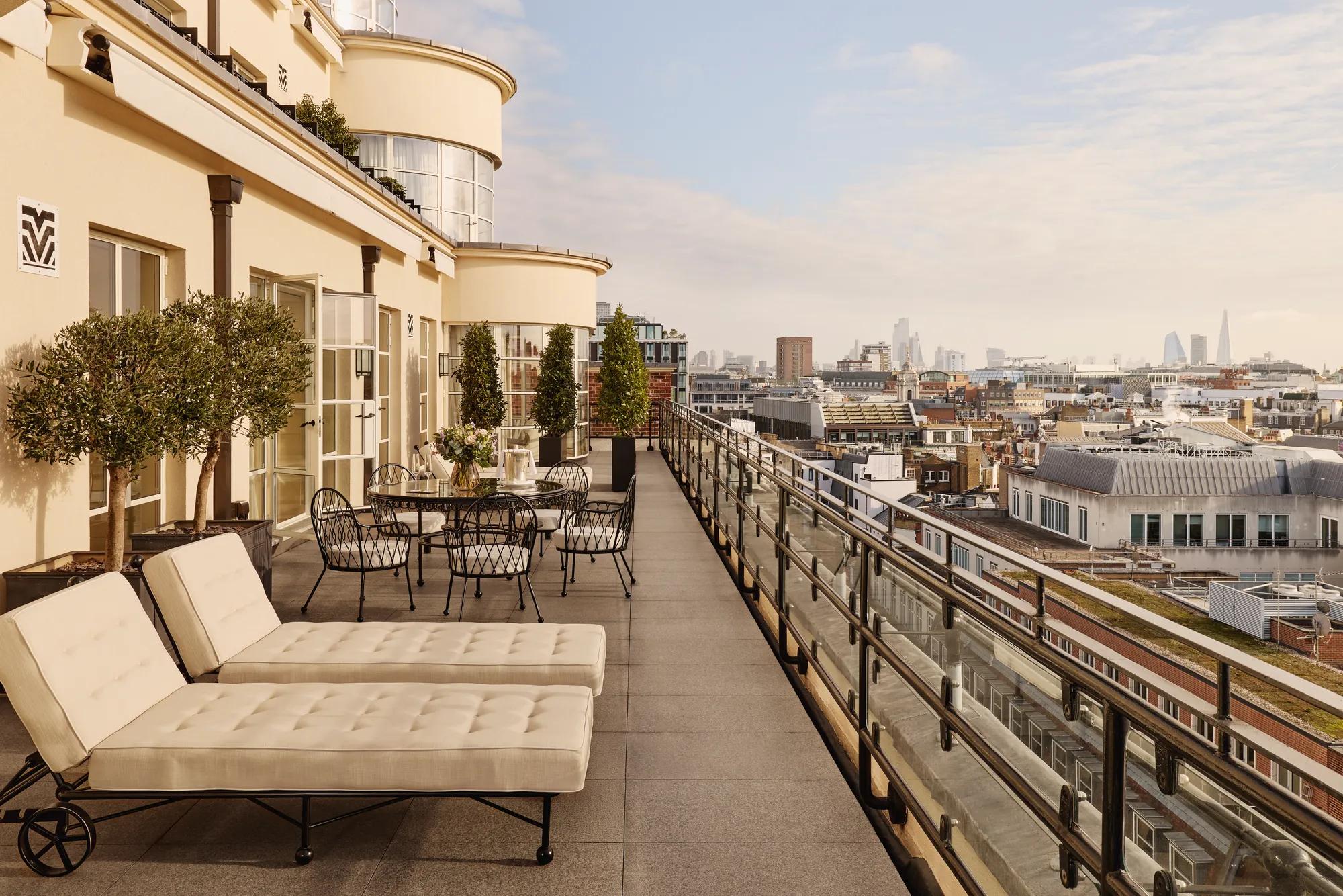 Expansive rooftop terrace with cushioned sun loungers, outdoor dining tables, potted trees, and glass railings, offering panoramic views of London’s skyline under a bright sky.