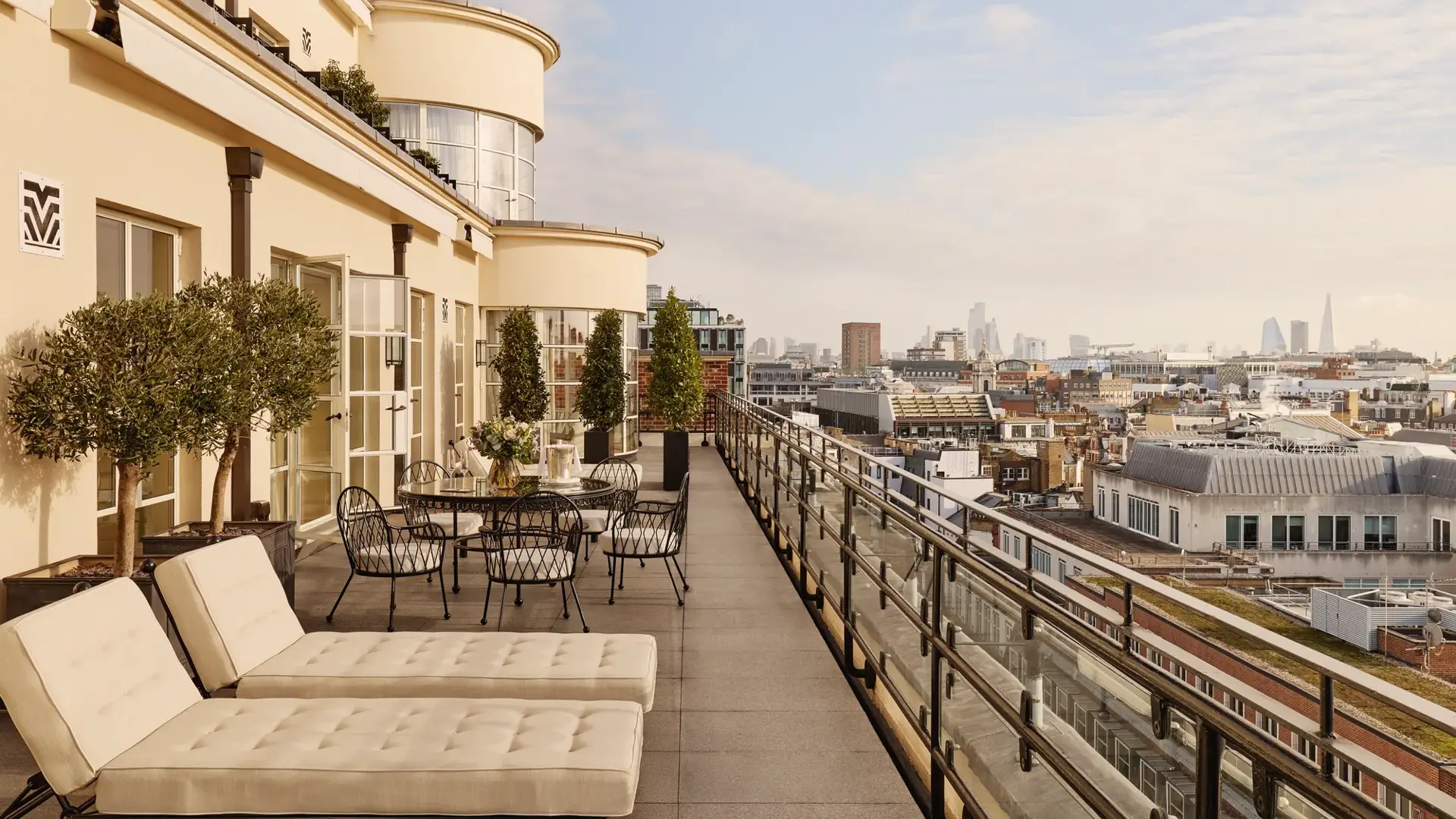 Expansive rooftop terrace with cushioned sun loungers, outdoor dining tables, potted trees, and glass railings, offering panoramic views of London’s skyline under a bright sky.