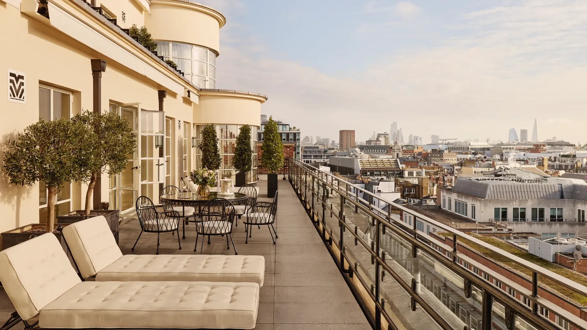 Expansive rooftop terrace with cushioned sun loungers, outdoor dining tables, potted trees, and glass railings, offering panoramic views of London’s skyline under a bright sky.