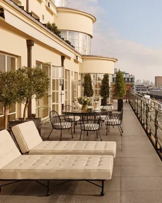 Expansive rooftop terrace with cushioned sun loungers, outdoor dining tables, potted trees, and glass railings, offering panoramic views of London’s skyline under a bright sky.