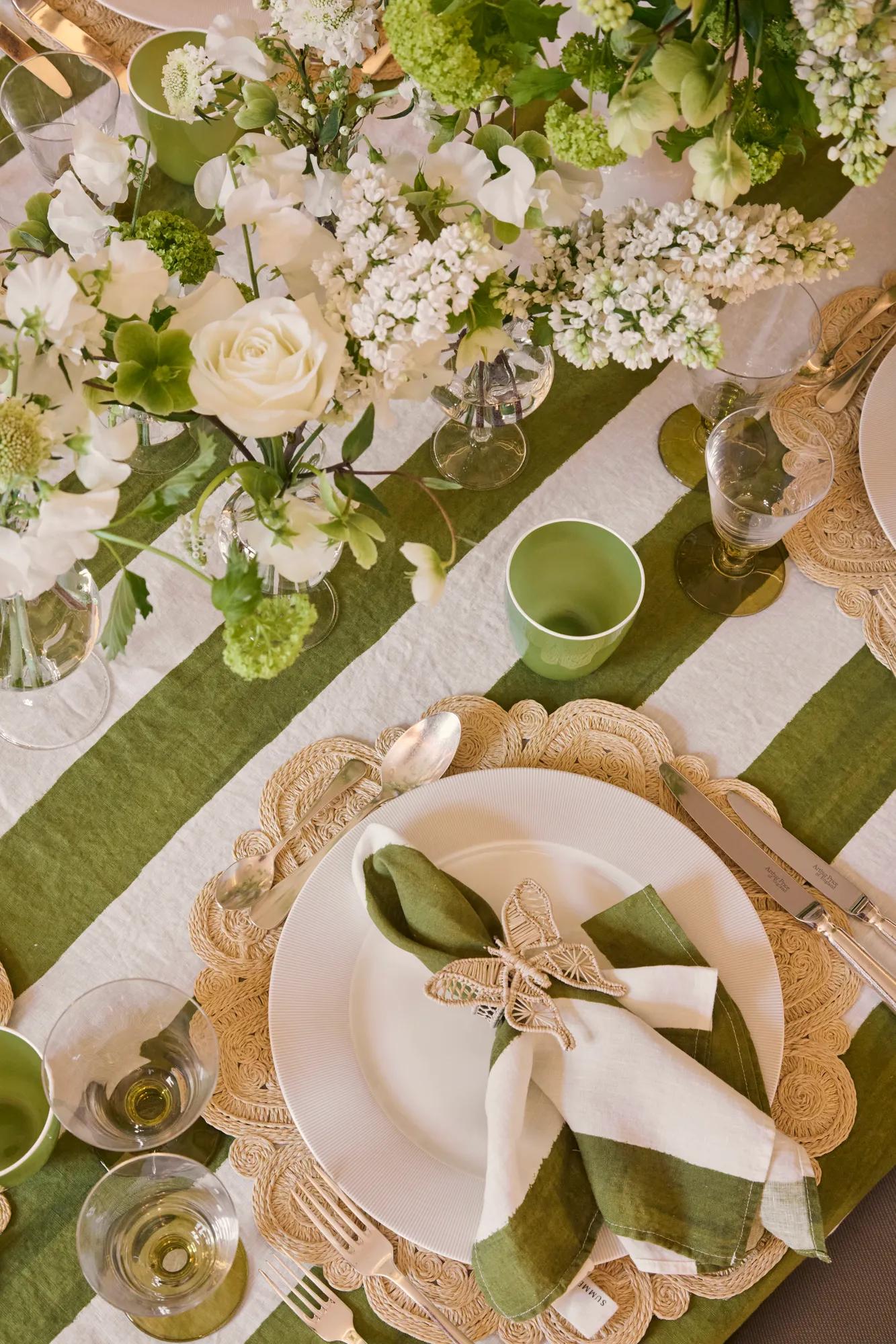 Overhead view of a dining table with striped green linens, white floral arrangements, woven placemats and folded napkins tied with butterfly decorations.