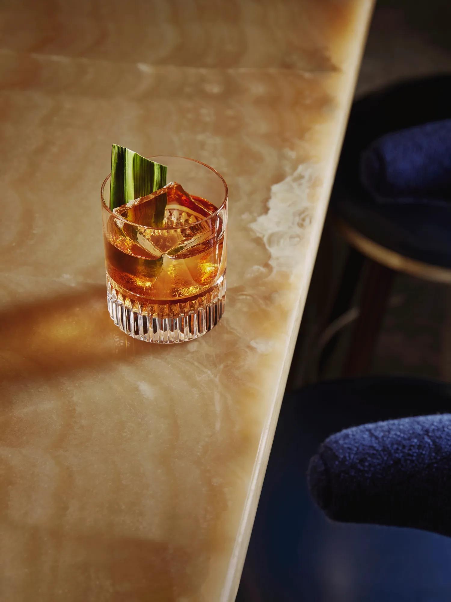 Old fashioned–style drink with large cube and green leaf garnish on the onyx bar edge.