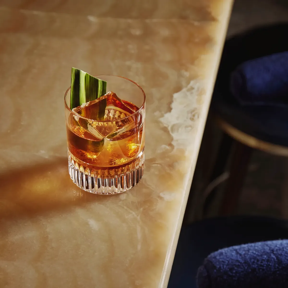 Old fashioned–style drink with large cube and green leaf garnish on the onyx bar edge.