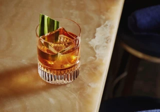 Old fashioned–style drink with large cube and green leaf garnish on the onyx bar edge.
