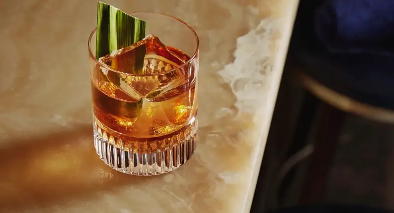 Old fashioned–style drink with large cube and green leaf garnish on the onyx bar edge.