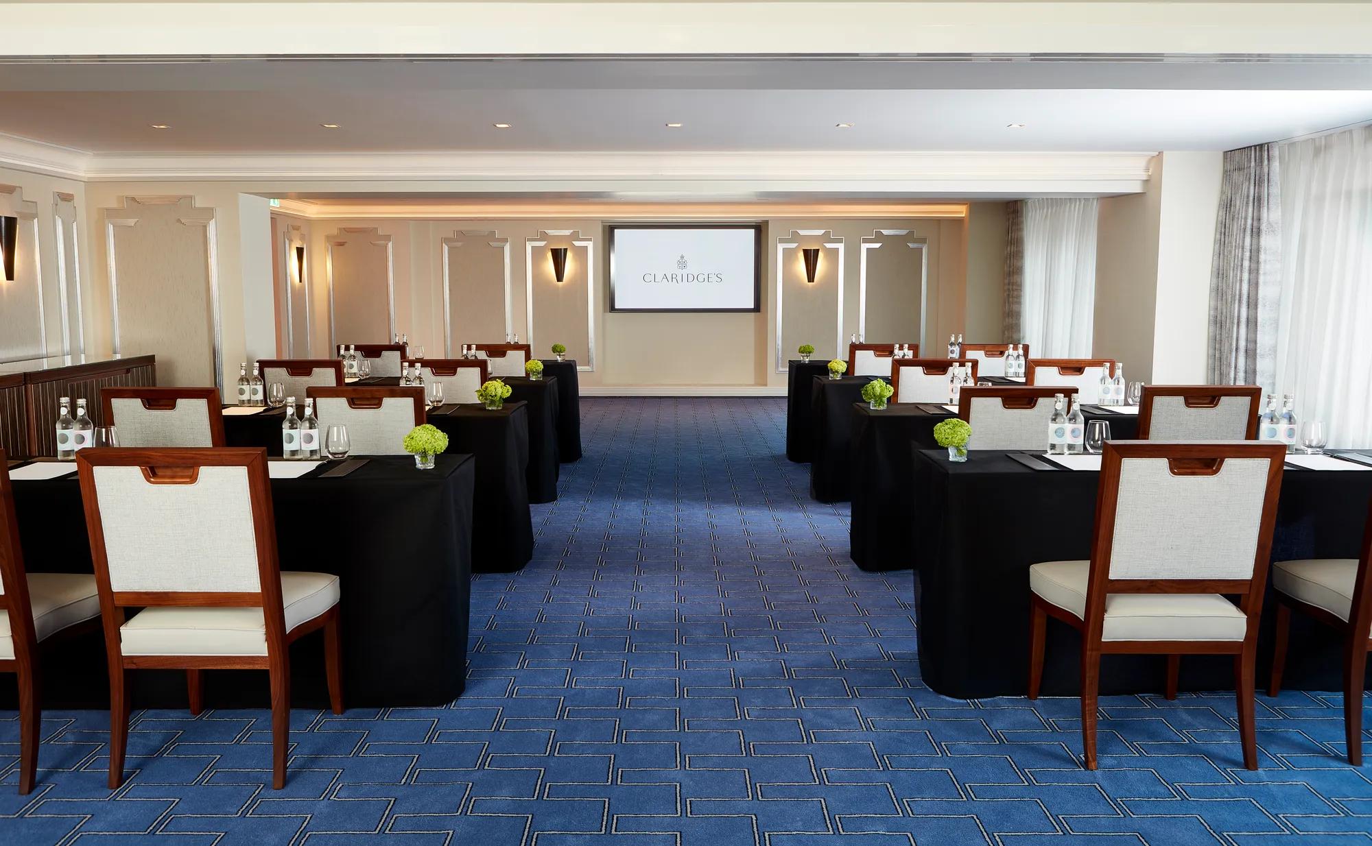 Modern meeting room at Claridge’s arranged in classroom style with dark tablecloths, wooden chairs, and bottled water, facing a presentation screen at the front of the room.