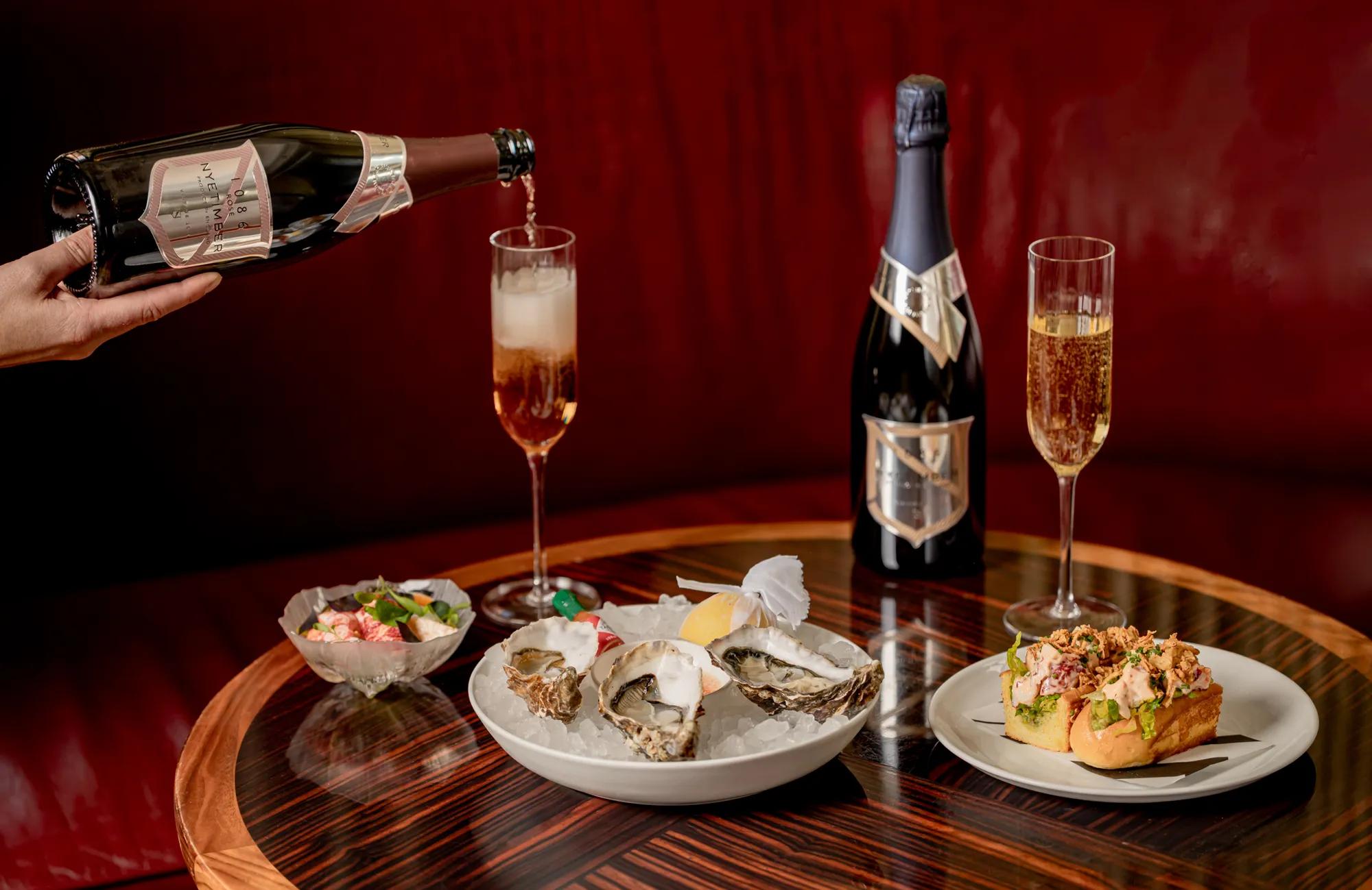 Hand pours sparkling wine into a flute beside oysters on ice, canapés and a champagne bottle on a polished table against a red banquette backdrop.