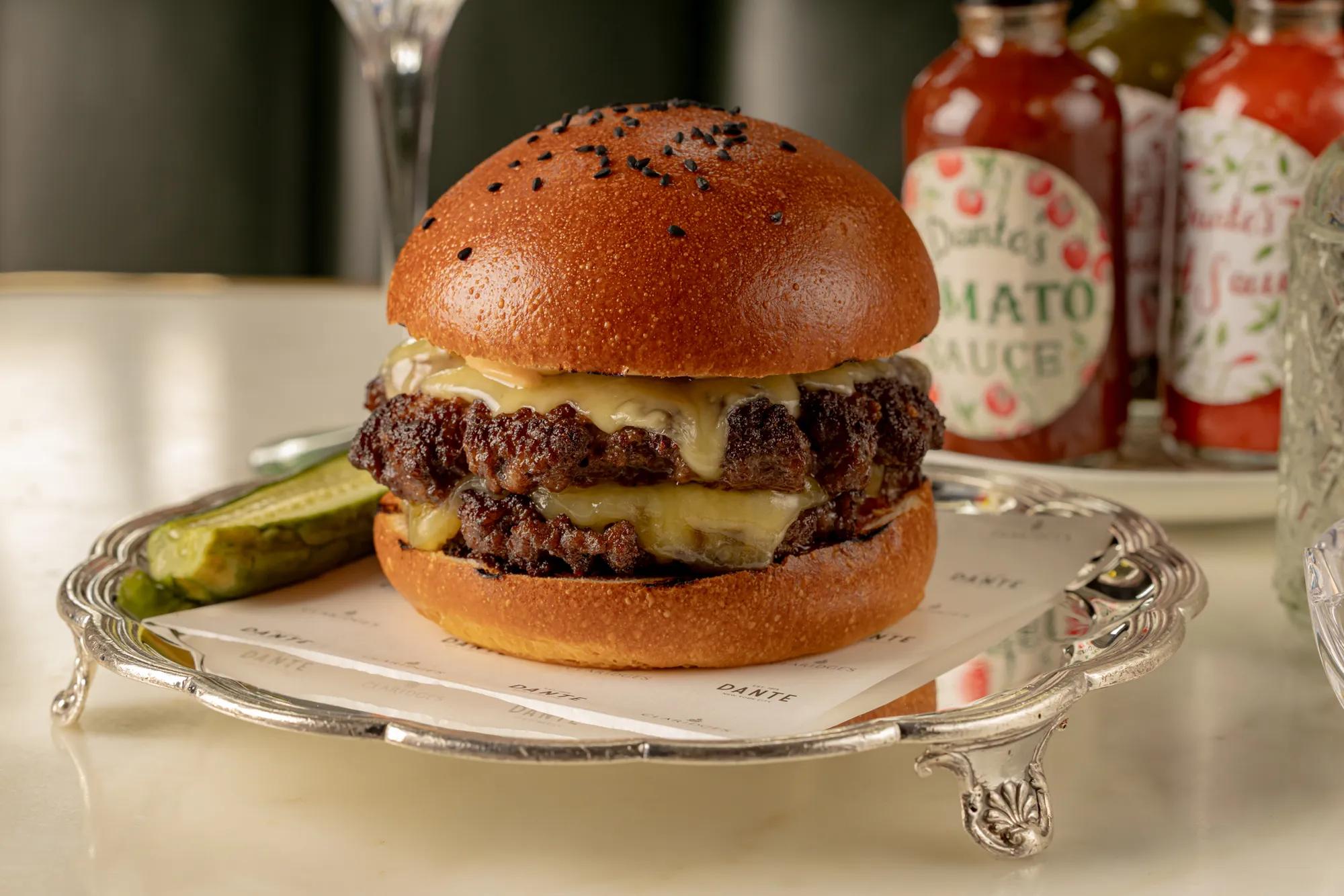 Double cheeseburger with melted cheese on a toasted brioche bun, served on a silver tray with pickle spears and bottles of sauce in the background.